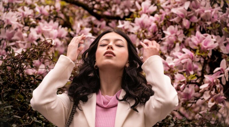 Woman stands among blossoming trees on a sunny spring day, illustrating the link between pollen season and hay fever skin.