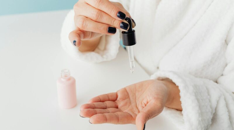 A person in a white dressing gown dispensing hydrating serum from a glass dropper onto the palm of their hand.