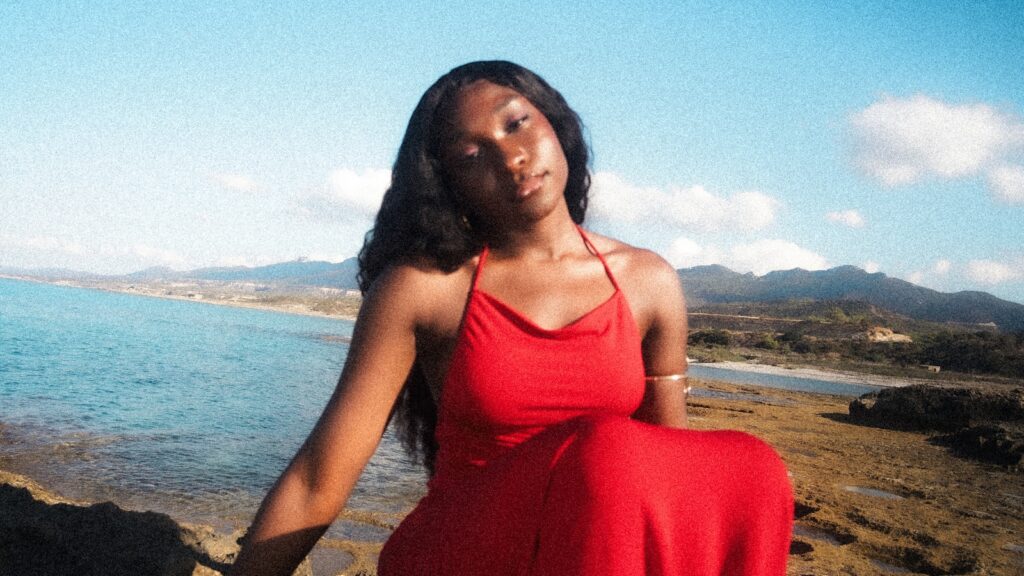 Editorial portrait of a woman in a flowing red dress by the sea, illustrating a summer holiday wardrobe UK 2026 capsule edit
