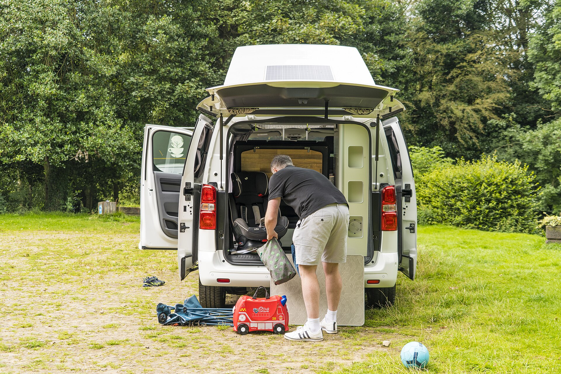 Parent packing a campervan with a child seat and ride-on suitcase before a summer holiday