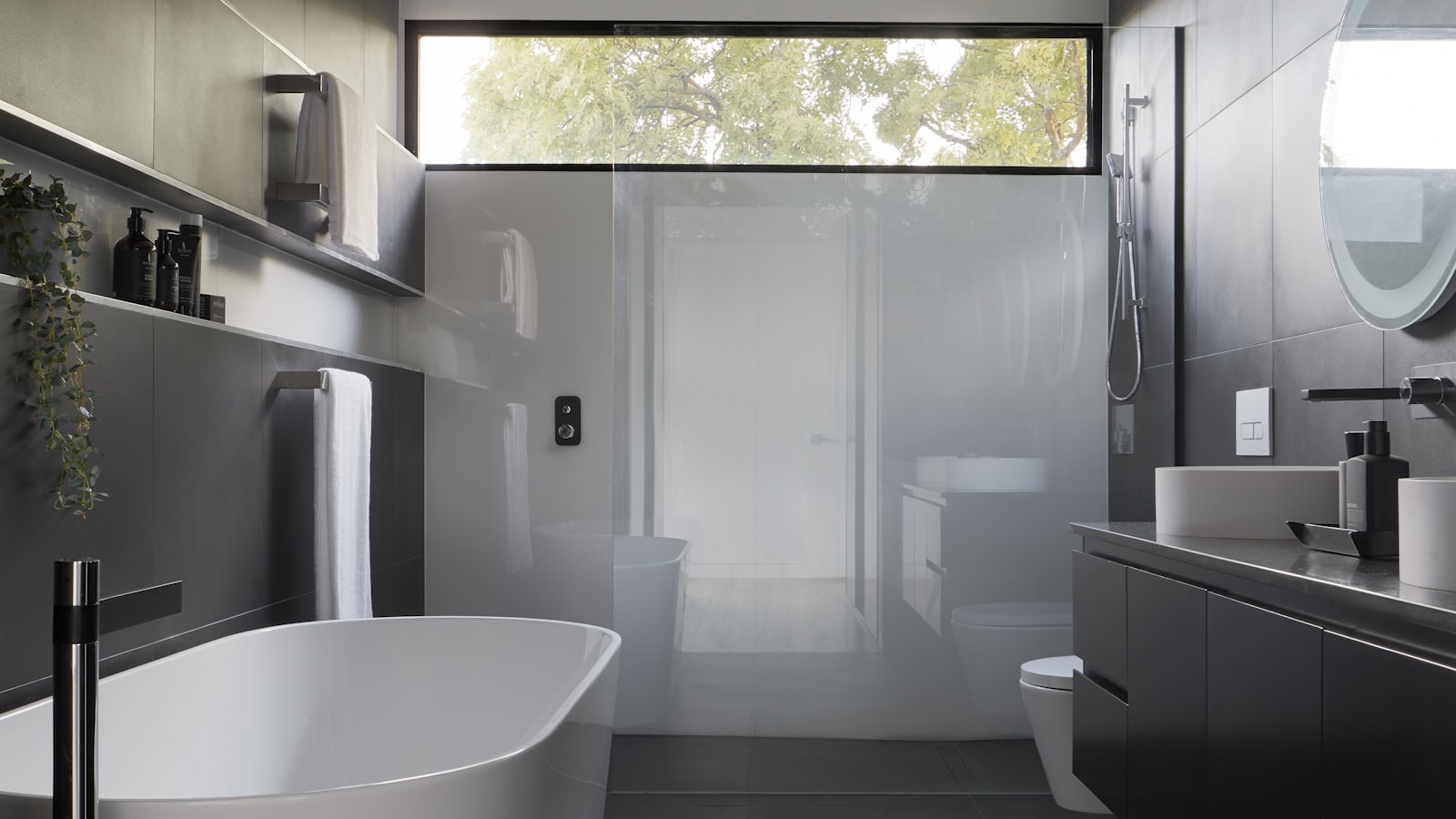 Dark tiled bathroom with a long recessed wall niche holding bottles above the bath