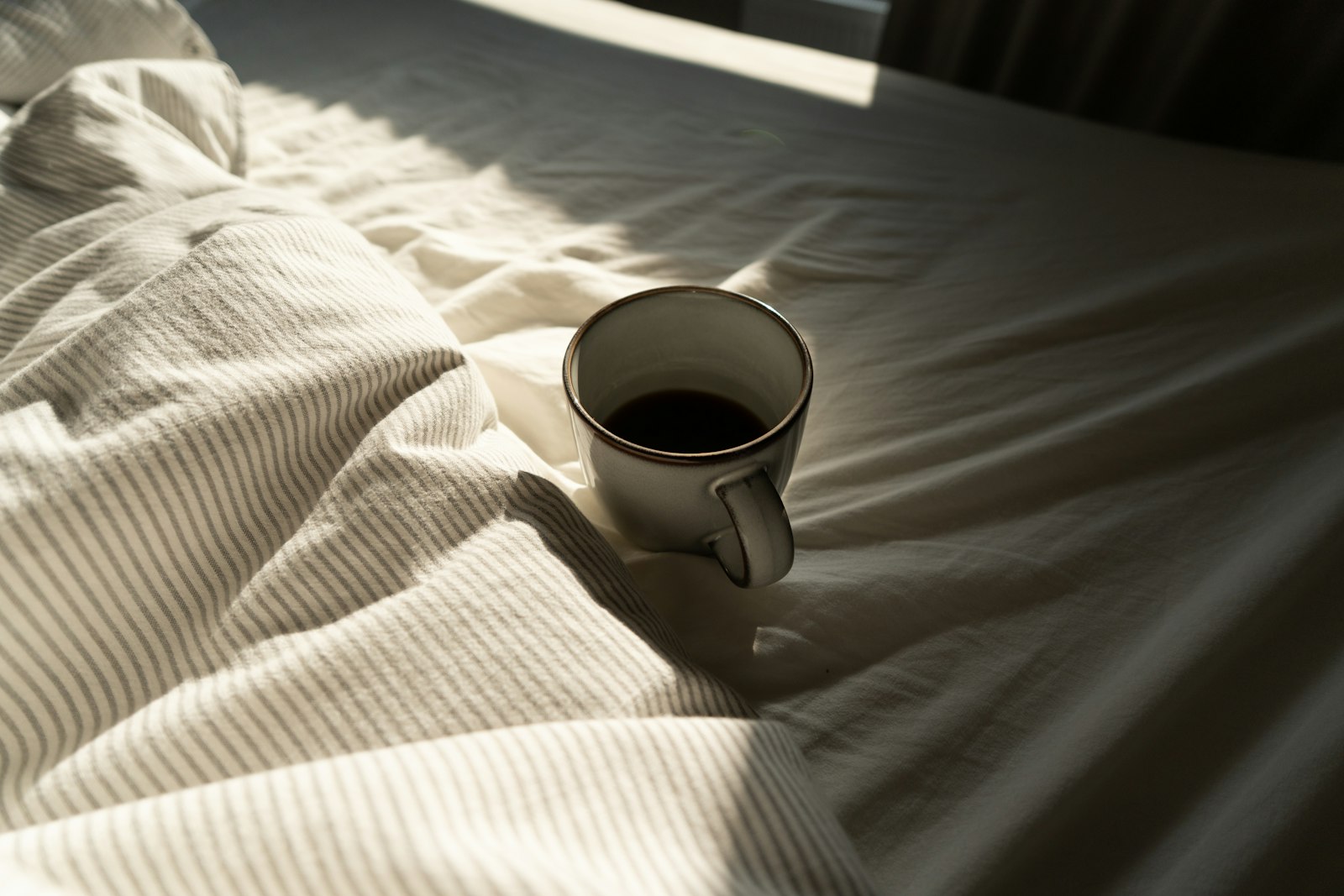 Mug of coffee resting on rumpled white bedding in morning light