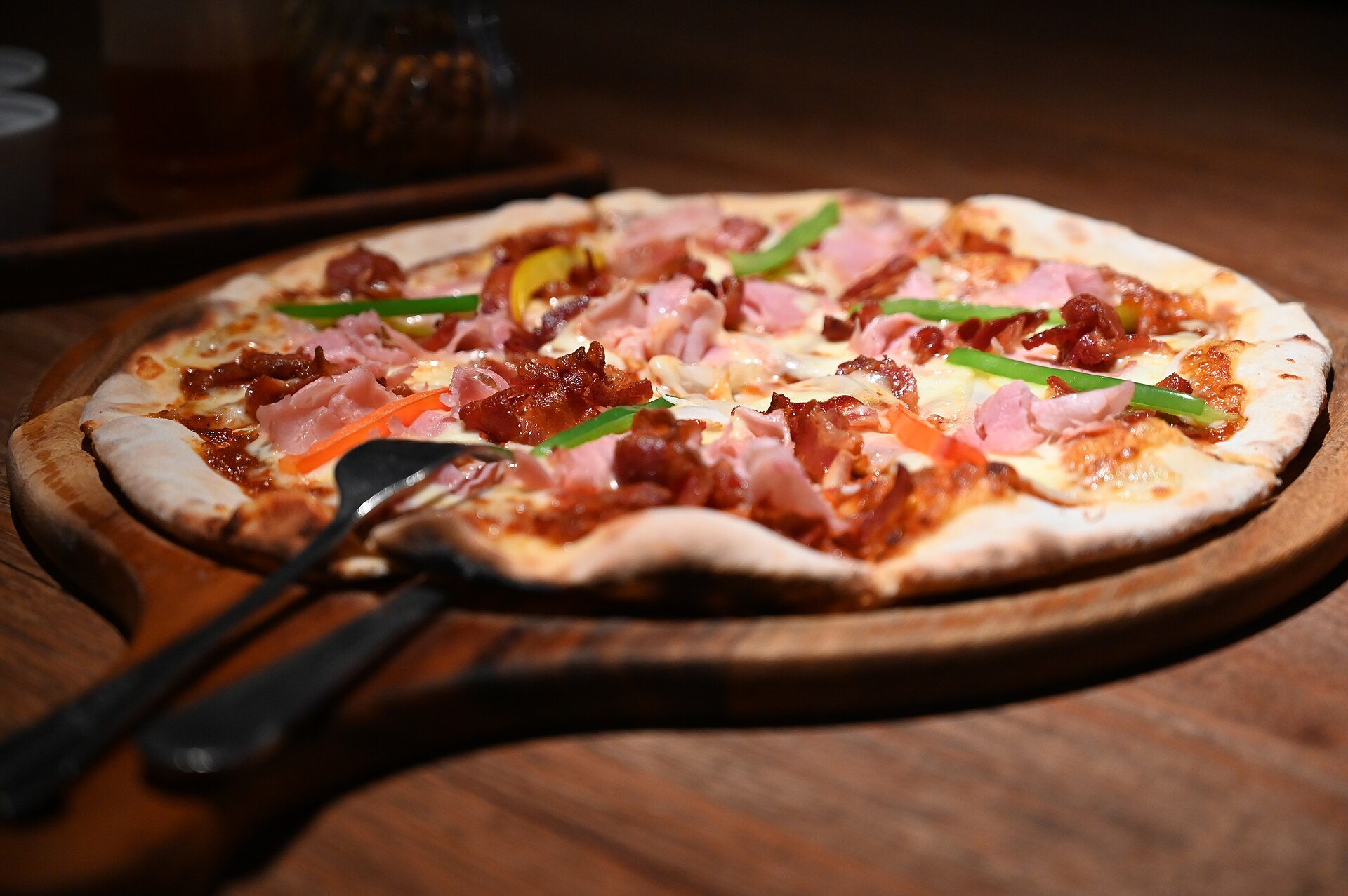 Thin-crust pizza on a wooden serving board with ham and peppers