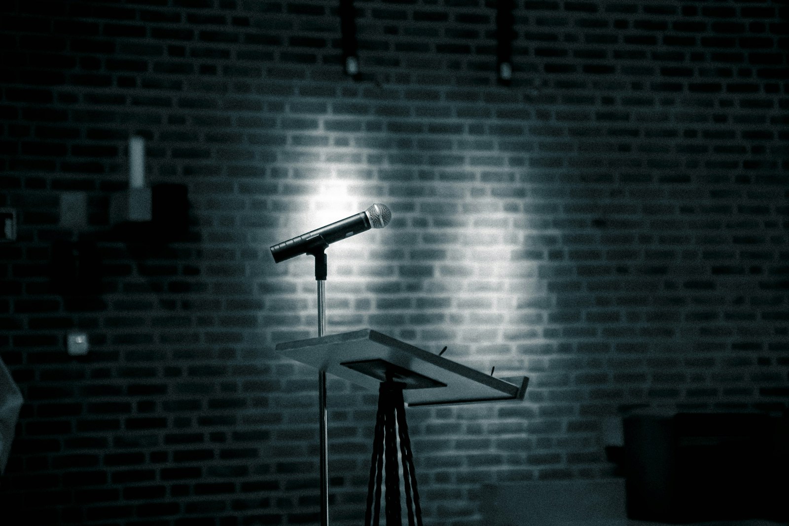 A microphone lit by a single spotlight against a brick wall in an empty comedy venue