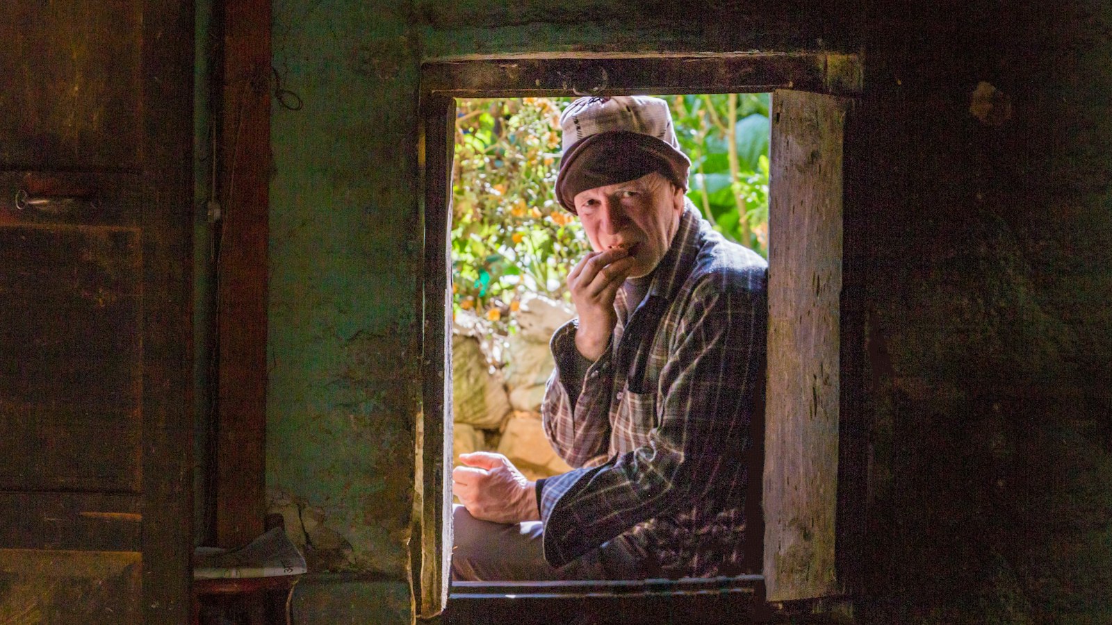 An elderly man in a flat cap looking thoughtfully out of a wooden window frame
