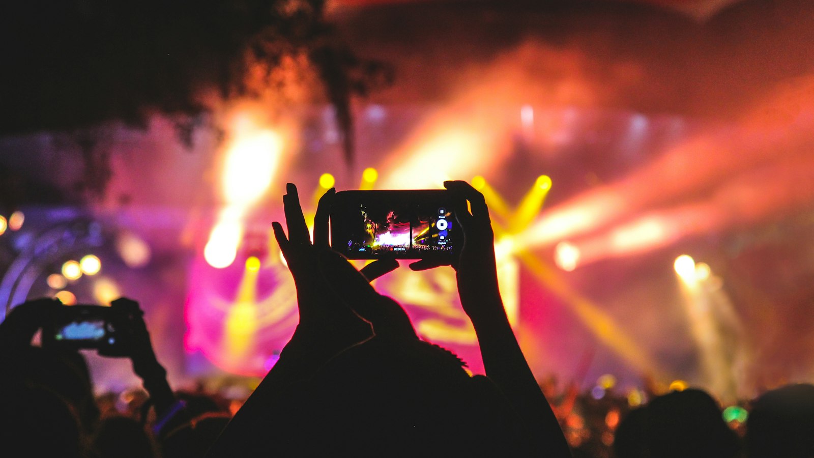 Fan filming an arena concert on a phone with stage lights behind, the kind of view a £55.60 Charli XCX UK tour 2027 ticket buys