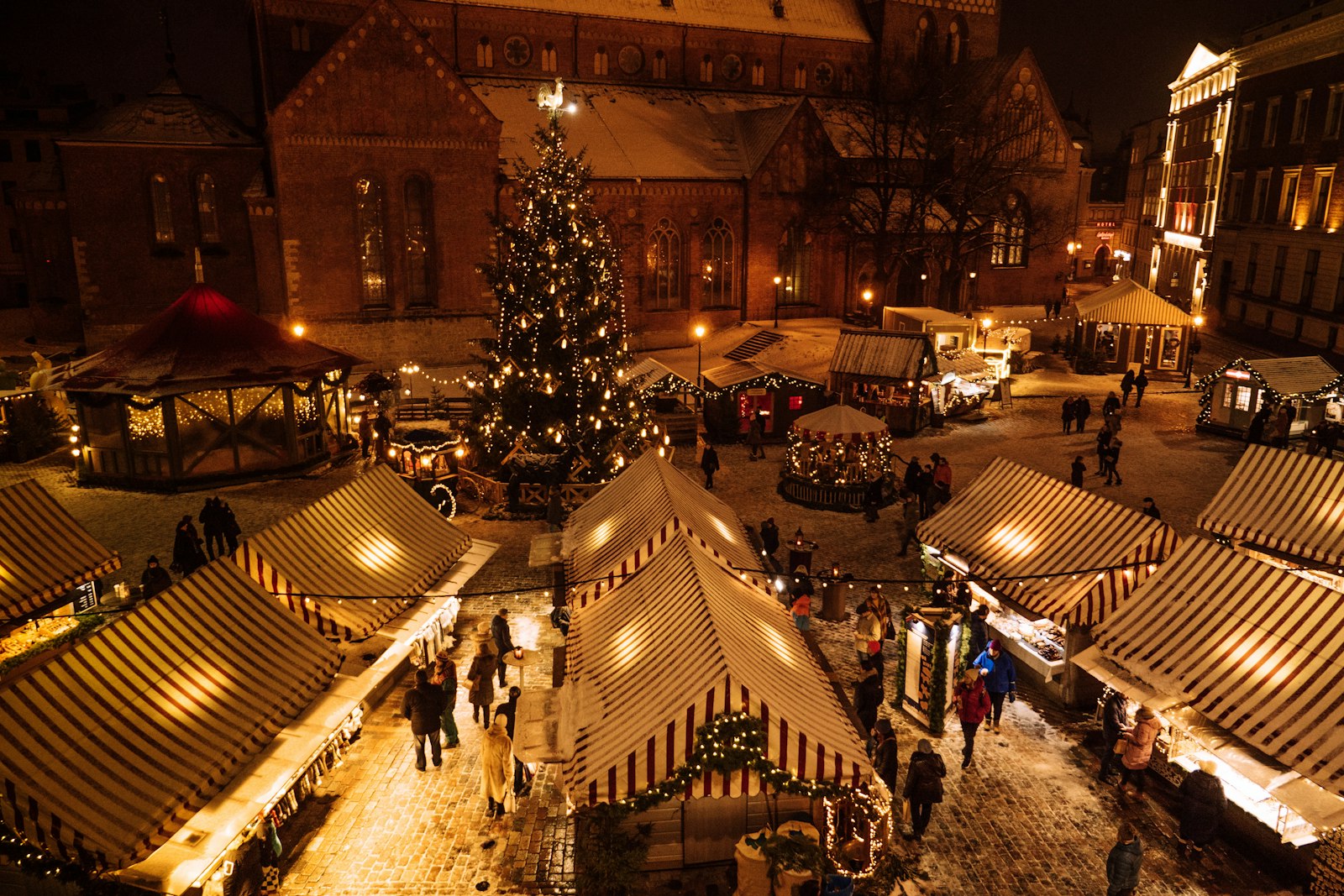 Aerial view of a European Christmas market square at night with a lit tree and striped stalls