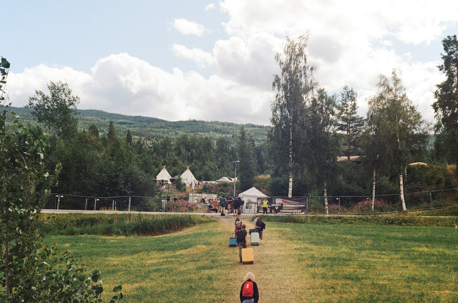 Festivalgoers carrying bags and camping gear across a grass field towards a festival entrance