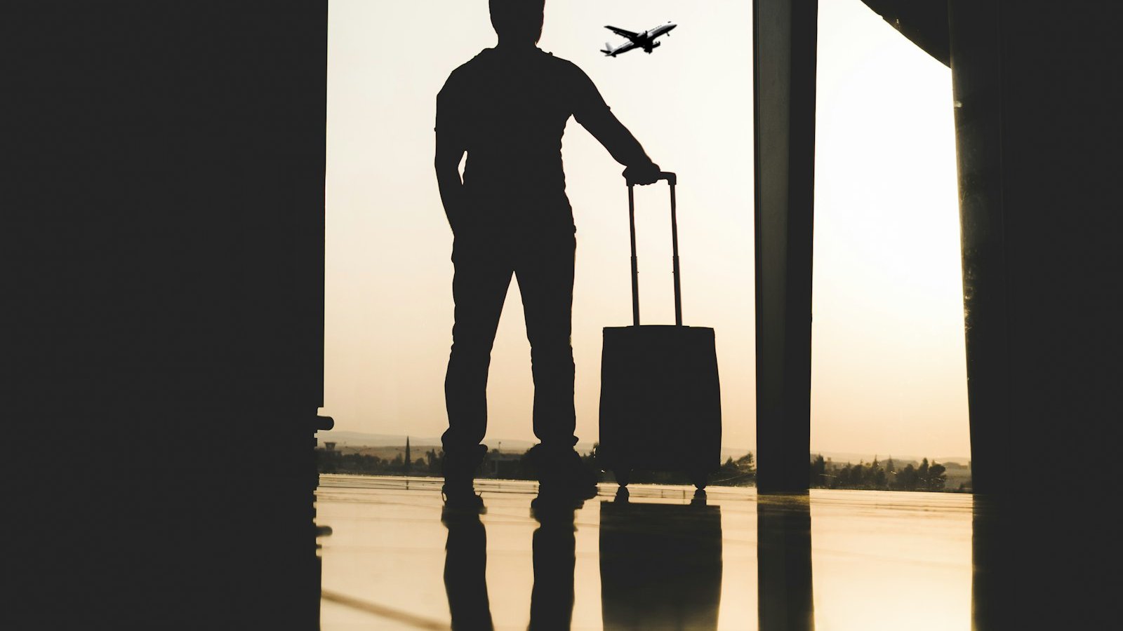 Silhouette of a traveller with a wheeled suitcase at an airport window as a plane takes off