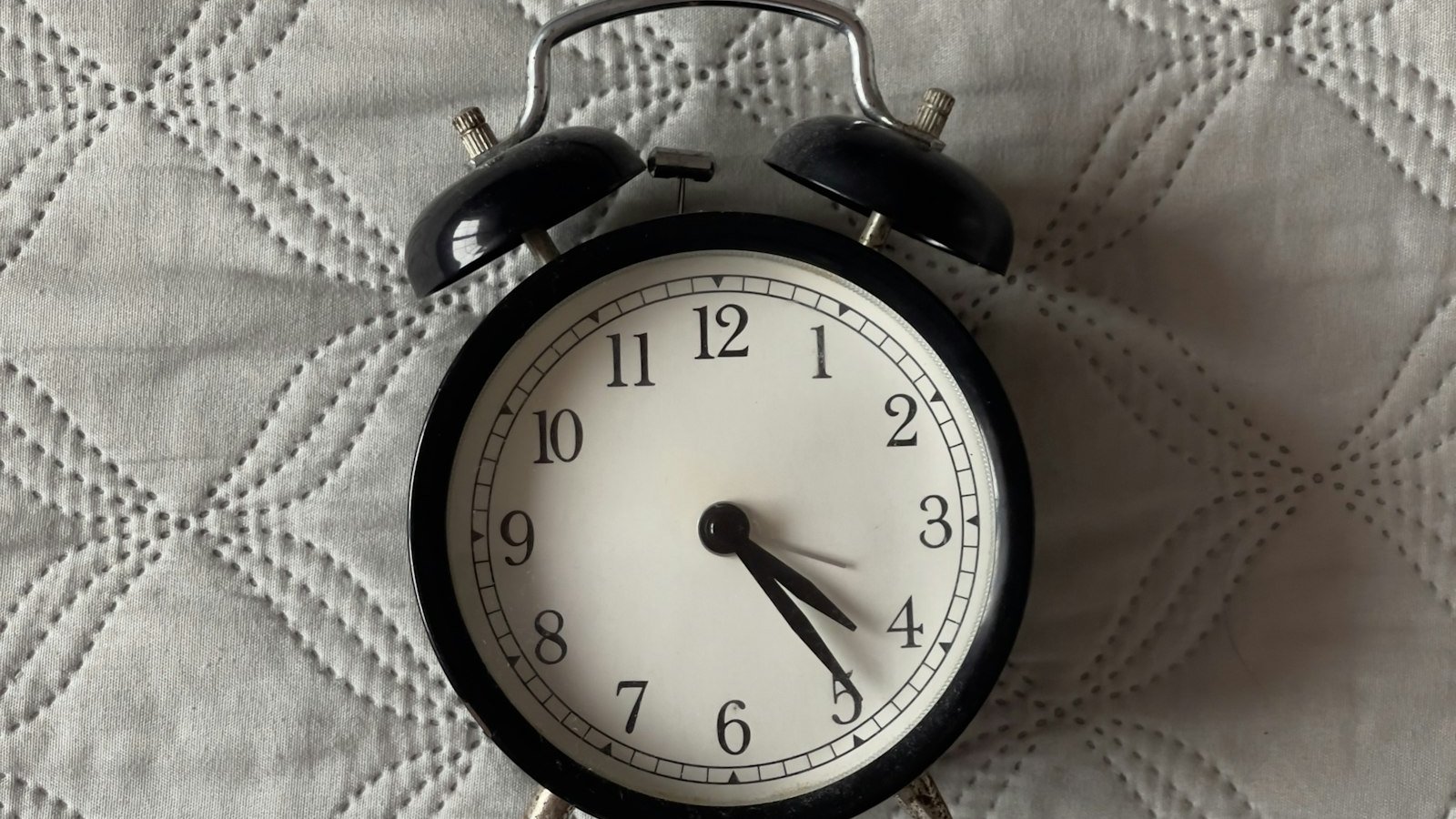 Black analogue alarm clock on a quilted bed, a reminder that your caffeine cut-off time is set hours before bedtime