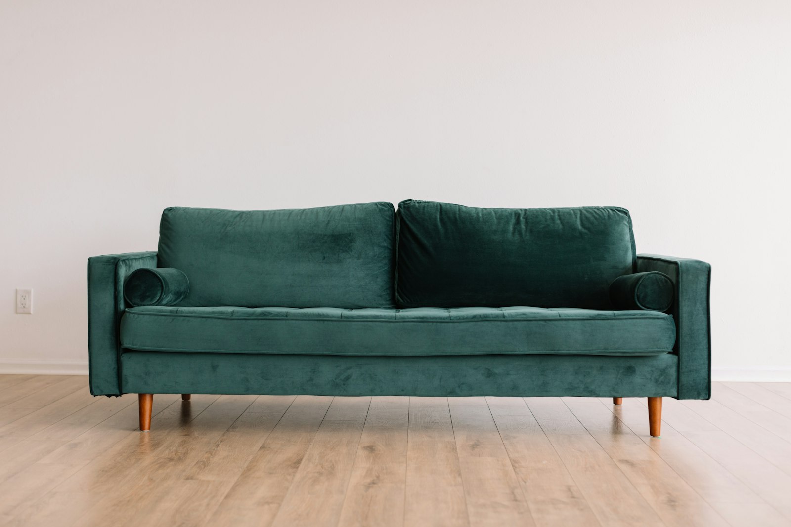 A deep green velvet sofa with tapered wooden legs against a white wall, showing how a bold seat anchors a room
