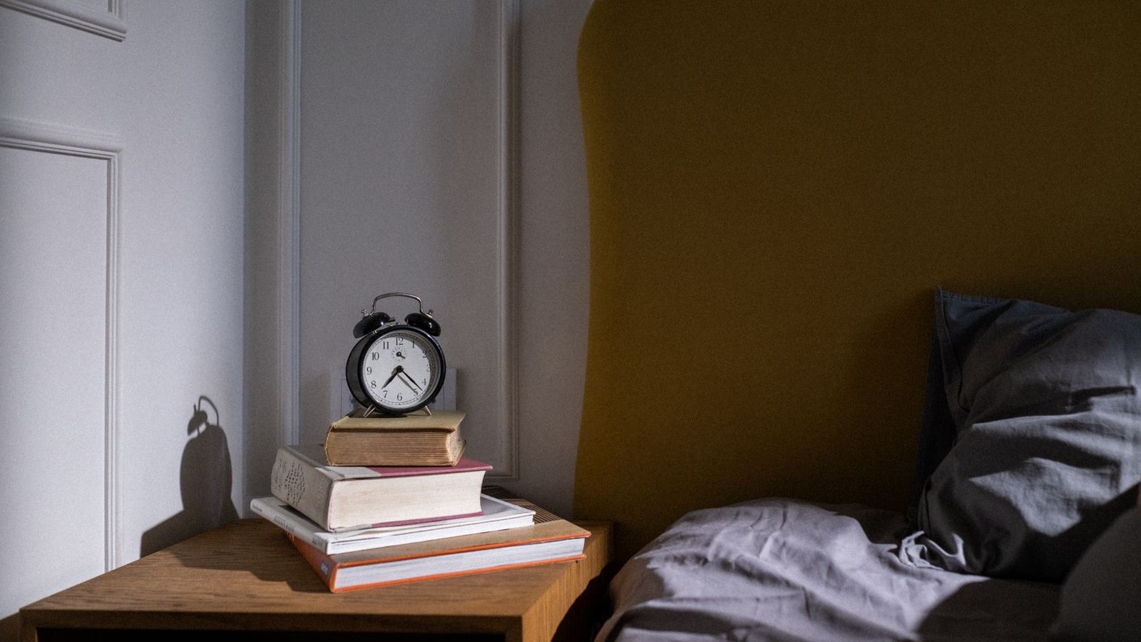 Black alarm clock on a stack of books beside a bed in low morning light